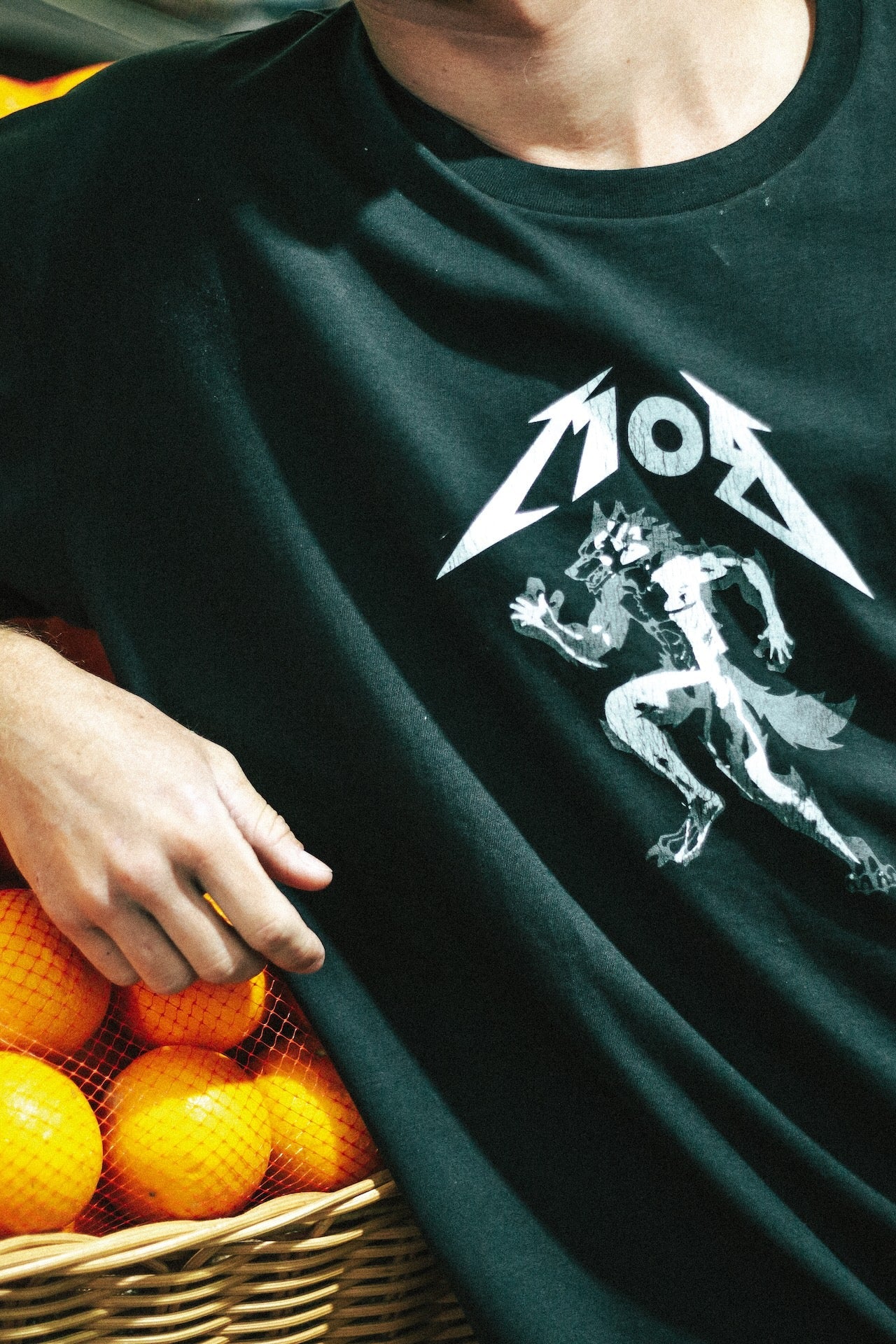 Person wearing a black t-shirt with a white graphic design, standing next to a basket of oranges.
